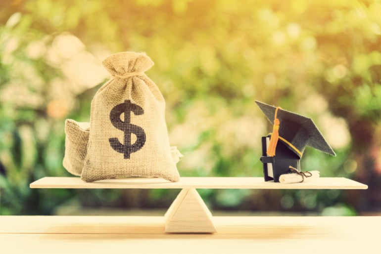 Money cost saving for goal and success in school, education concept : US dollar bills / cash in hessian bags, a black graduation cap or hat, a certificate / diploma and a book on simple balance scale. shutterstock_1091618918