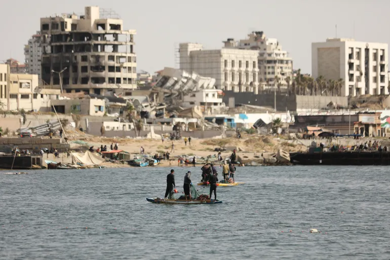 Palestinian fishermen try to fish with rowing boats and fishing rods on the coast of Gaza city as damaged and destroyed buildings are seen behind in Gaza غيتي