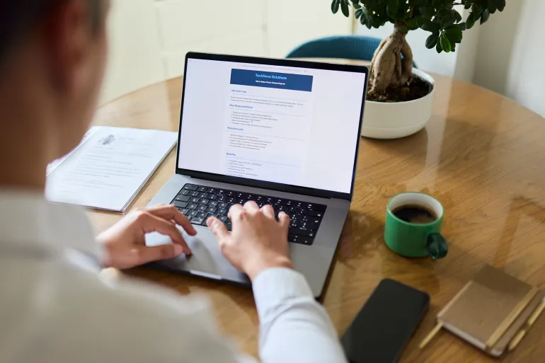 Man sitting at home in front of his laptop browsing job listings. A printed copy of his resume is placed beside the tablet on the table. He appears focused on the job opportunity, surrounded by a simple, professional office setting. The image represents job search, remote work, and digital communication.