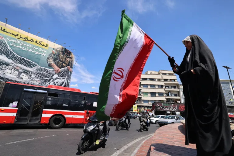 A woman waves Iran's national flag in front of a giant billboard reading 'The Strait of Hormuz remains closed' at the Revolution Square in Tehran on April 12, 2026.