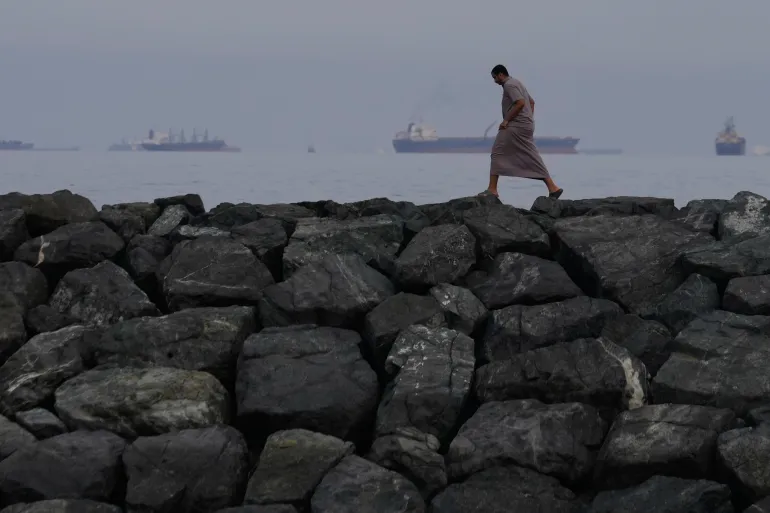 FILE- A man walks along the shore as oil tankers and cargo ships line up in the Strait of Hormuz, seen from Khor Fakkan, United Arab Emirates, March 11, 2026. (AP Photo/Altaf Qadri, File)