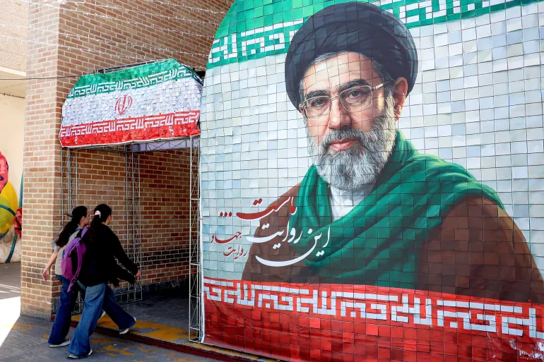 TOPSHOT - Women walk past a banner depicting Iran's current supreme leader Ayatollah Mojtaba Khamenei along a street Tehran on May 6, 2026.