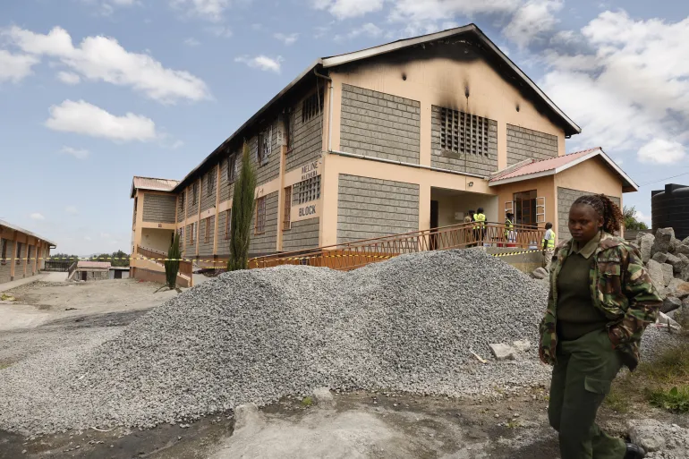 CORRECTION / TOPSHOT - A Kenyan police officer walks past forensic experts standing near the entrance of a building at Utumishi Girls Academy where a deadly fire took place in Gilgil, north of the capital Nairobi on May 28, 2026.