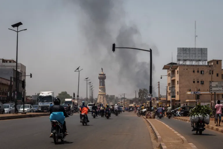 A column of black smoke rises above buildings as traffic passes the Africa Tower monument in Bamako on April 26, 2026. April 25, 2026's shock attacks, synchronised by Tuareg rebels of the Azawad Liberation Front (FLA) coalition and the jihadist Group for the Support of Islam and Muslims (JNIM), targeted several areas in the vast arid country. Fighting resumed on April 26 in several areas, including Kita near Bamako, Kidal, Gao and Severe. Tuareg rebels meanwhile announced an agreement allowing Russian forces backing Mali's army to withdraw from the northern city of Kidal, which they claimed was "totally" under their control. (Photo by AFP)