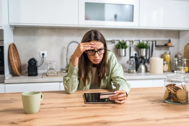 Anxious young woman deciding whether to pay online—holding a credit card and staring at a tablet concerned about fraud, hidden fees, and tight budgeting while shopping from home