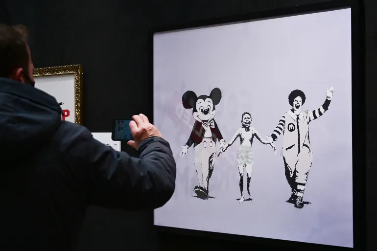 A man takes a picture of British street artist Banksy's mural "Napalm (Can't Beat that Feeling)" during a preview of the exhibition "The world of Banksy, the immersive experience" at the Milano Centrale main railway station in Milan on December 02, 2021. (Photo by MIGUEL MEDINA / AFP) / RESTRICTED TO EDITORIAL USE - MANDATORY MENTION OF THE ARTIST UPON PUBLICATION - TO ILLUSTRATE THE EVENT AS SPECIFIED IN THE CAPTION
