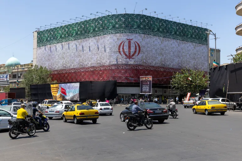 TEHRAN, IRAN - JUNE 15: Cars and monocycles are seen traveling in front of a billboard displaying the Iranian flag on June 15, 2026 in Tehran, Iran. According to reports, the U.S. and Iran have signed a preliminary agreement to end the war and open the Strait of Hormuz. (Photo by Majid Saeedi/Getty Images)