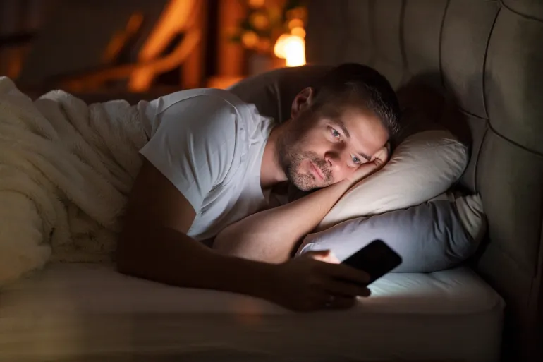 A sleepless man lying in dark bedroom with cellphone in hands and doom scrolling on social media.